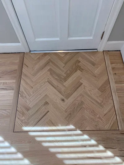 Herringbone pattern hardwood flooring installation at entryway with white door frame